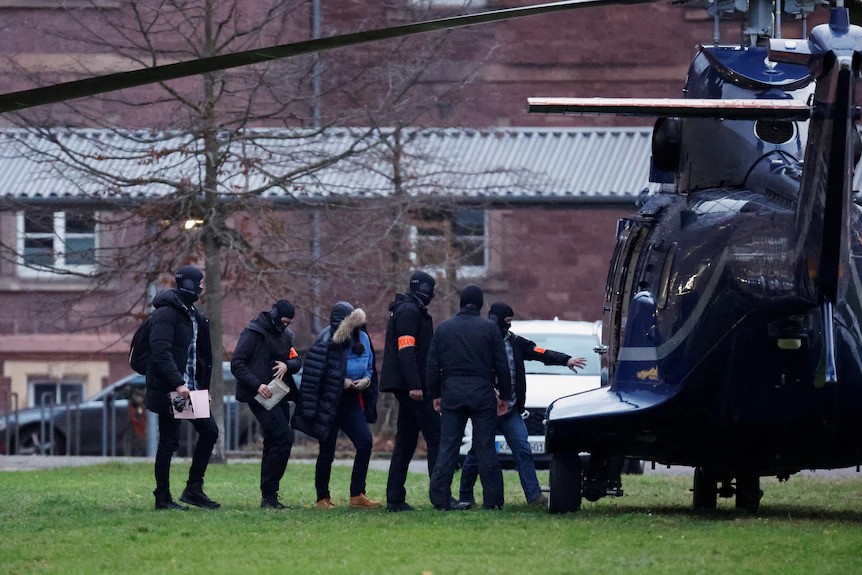 Police lead people into a helicopter outside a brick building. 