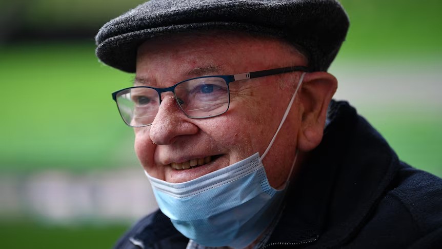 A close-up of an elderly man with glasses, a small cap and a face mask around his chin