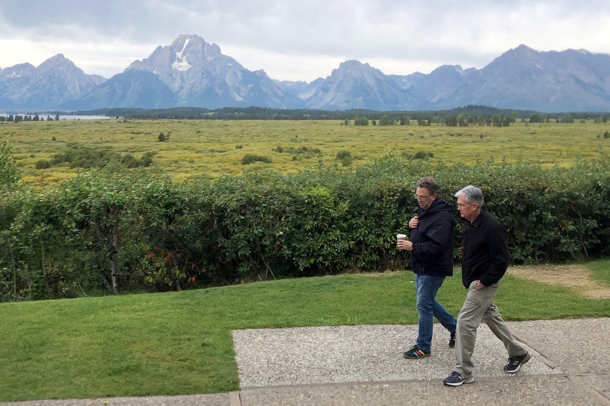 Two casually dressed men walk along a path with green fields and mountain ranges in the background.