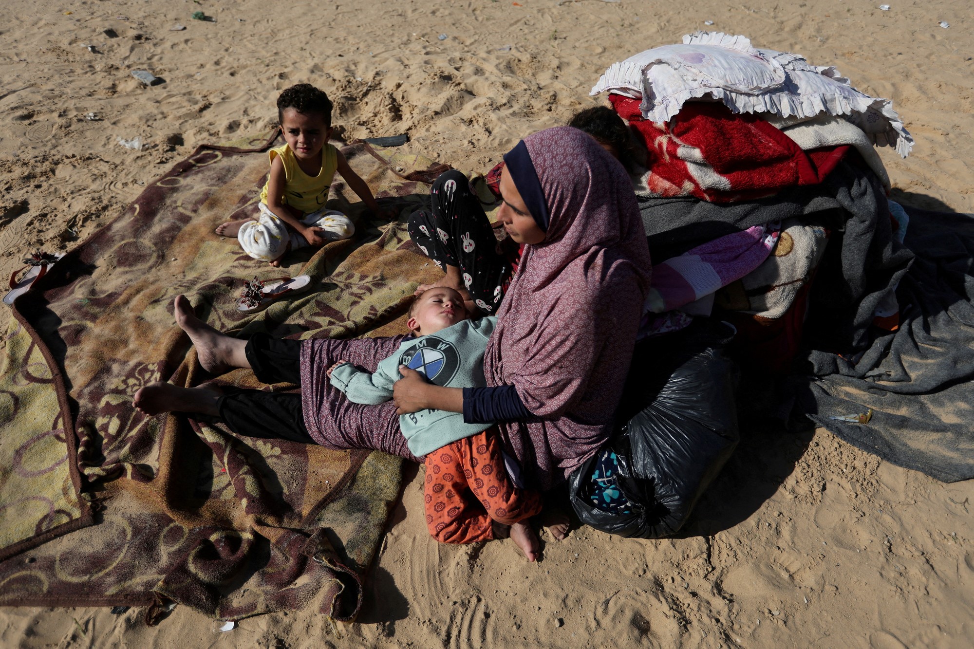 Woman sits with two children on sand with a bundle fof towels and bedding beside her. 