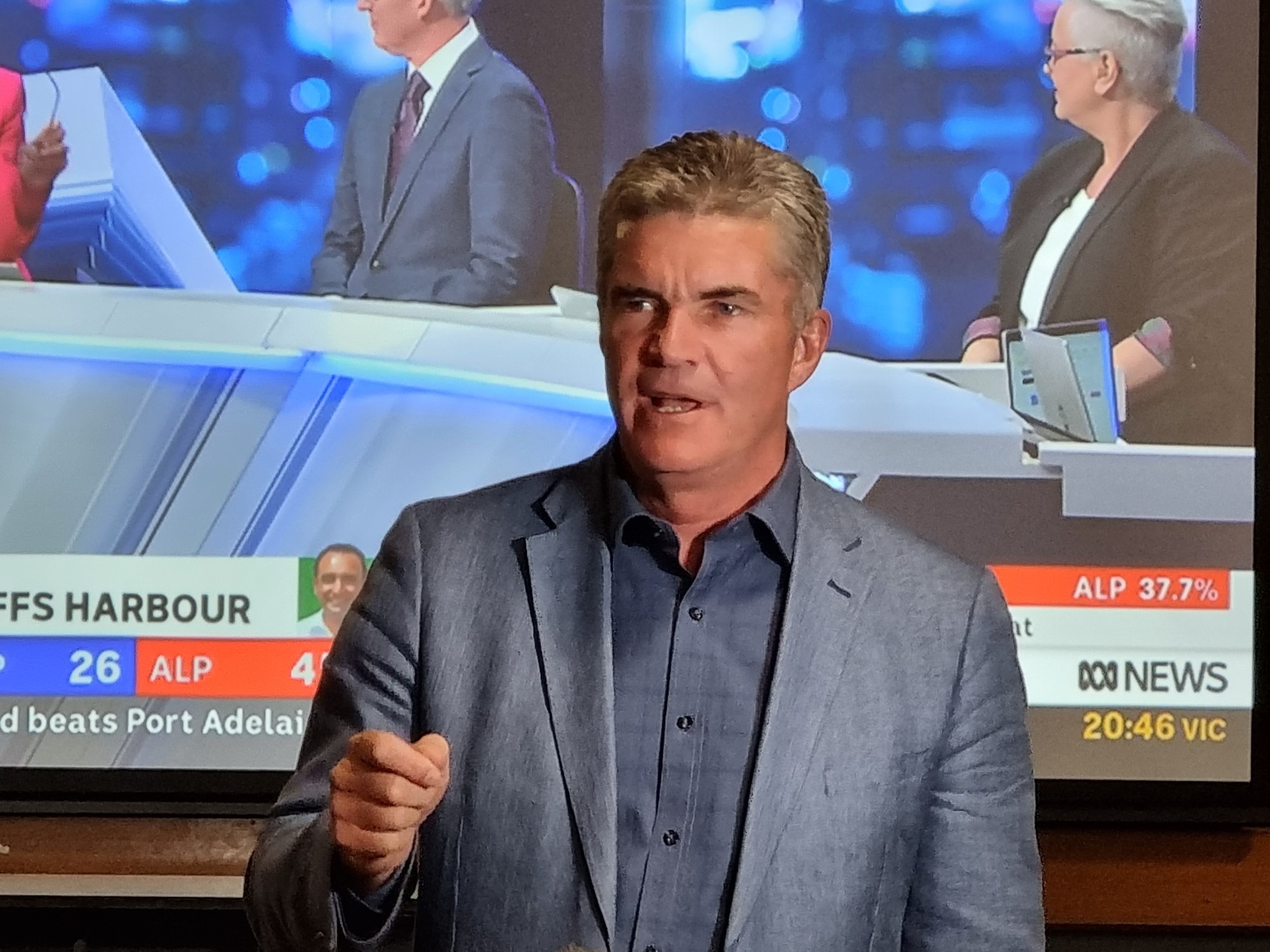 A man makes an emphatic fist while speaking in front of a big screen