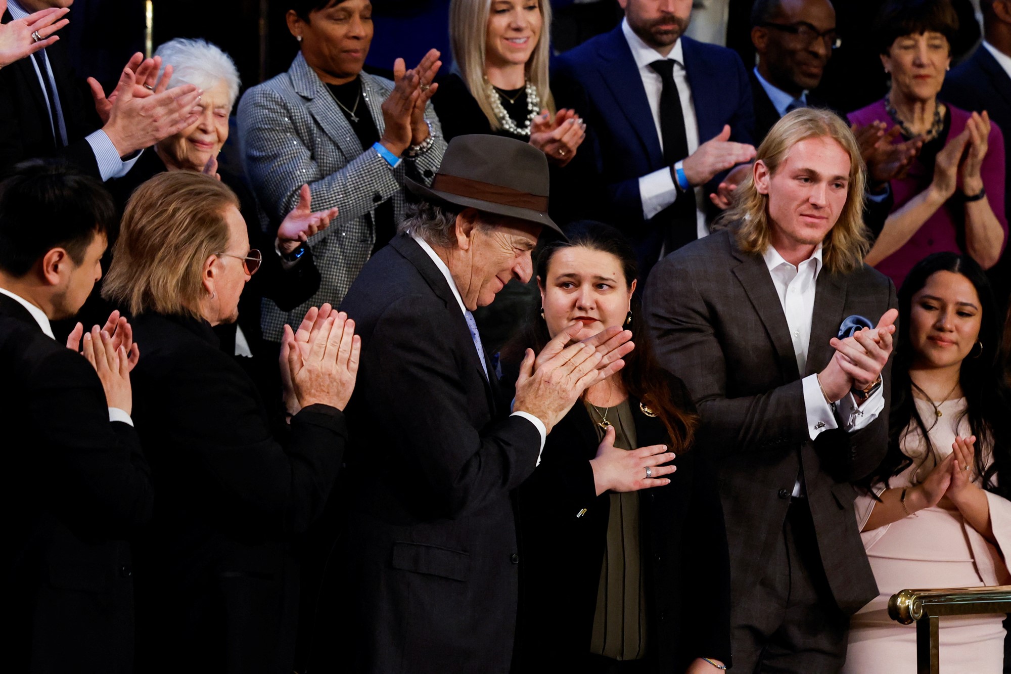 A man in a suit and hat smiles as he claps in a crowd of people in suits.