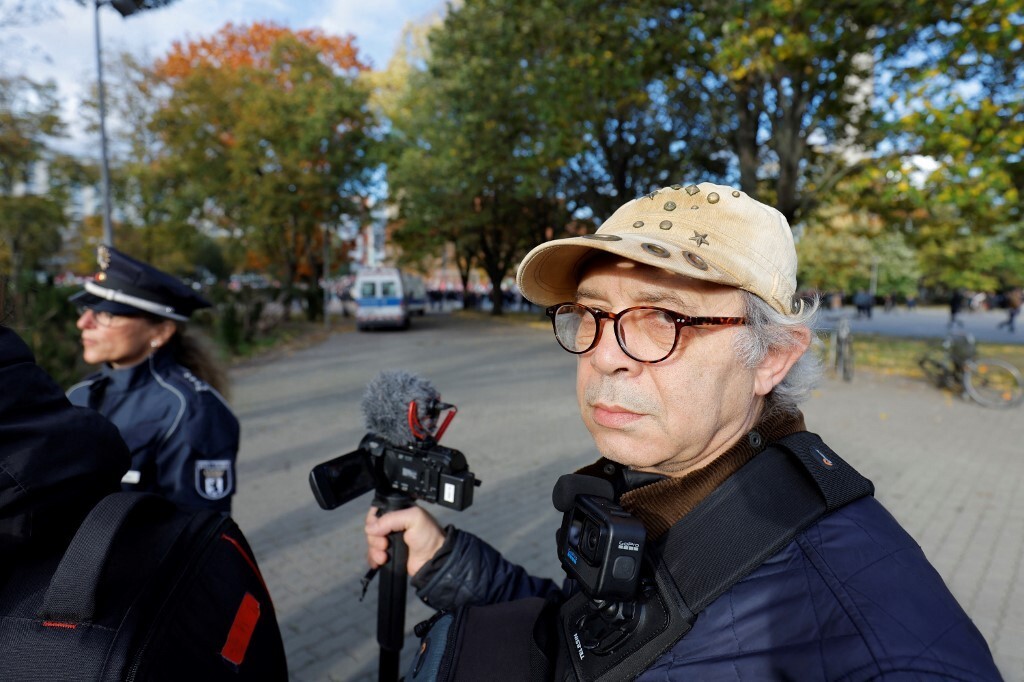 Man with glasses and cap holds tripod and camera in right hand. 