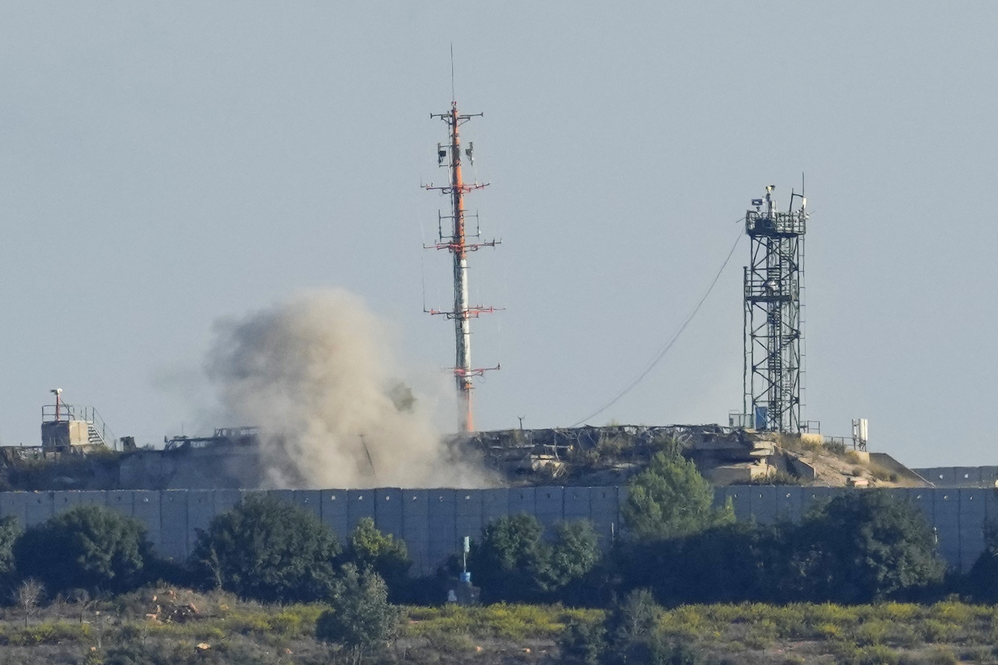 Smoke rises behind a large border wall, near an army base