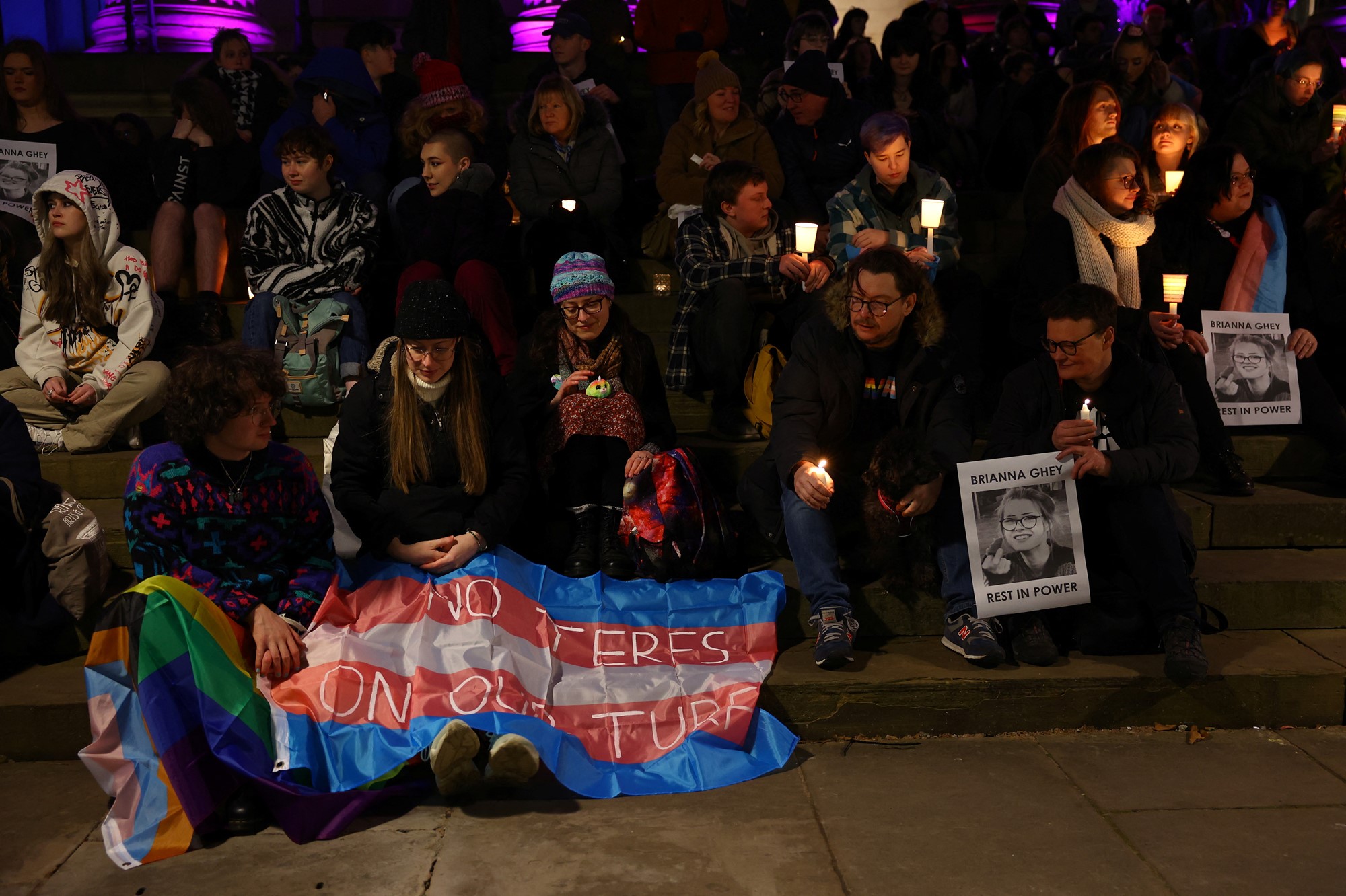 People hold candles, the trans flag with no terfs on our turf written on it, and photos of Brianna Ghey at her vigil.
