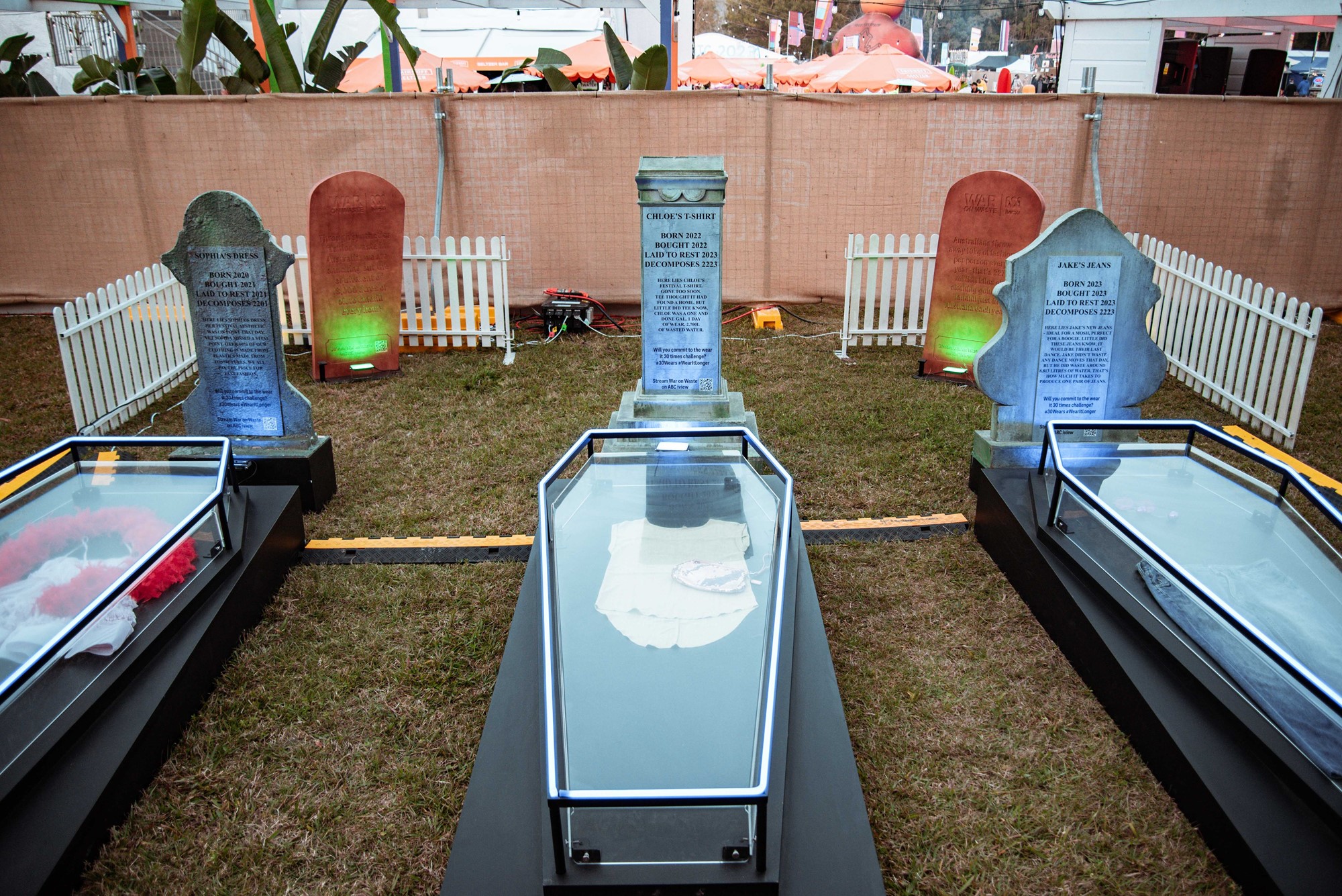 Three fake glass coffins with headstones that have a piece of fashion in them each