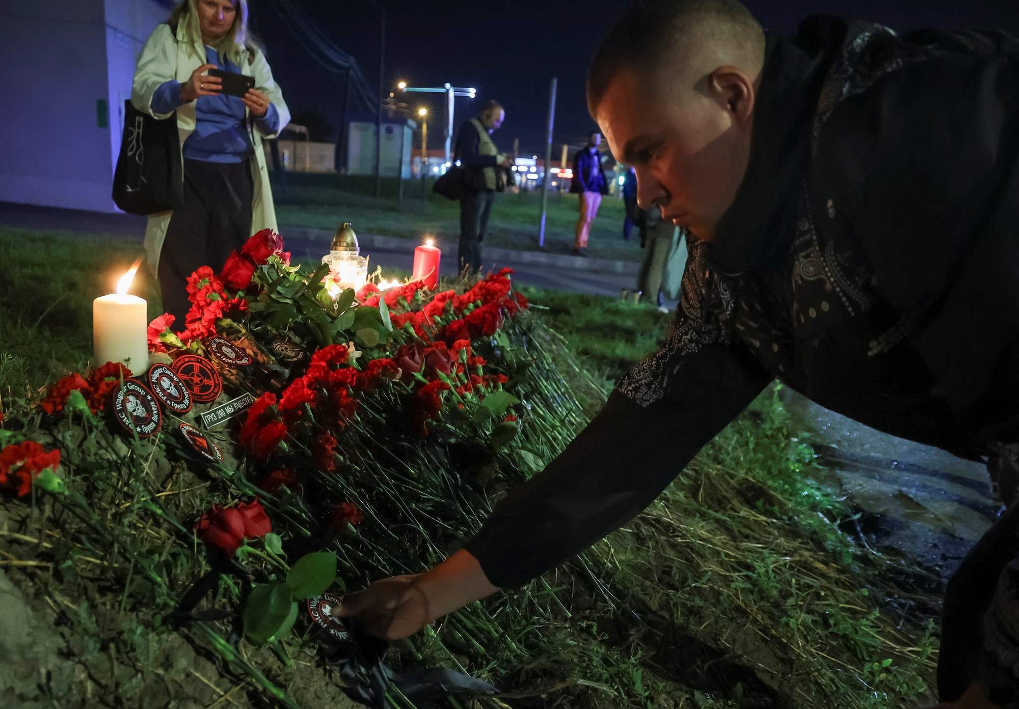 A man leans down to put a patch on a pile of roses, with candles, as a woman nearby takes a photo