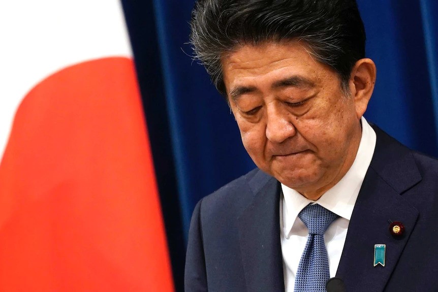 Shinzo Abe frowns as he looks down slightly, standing in a suit in front of a Japanese flag