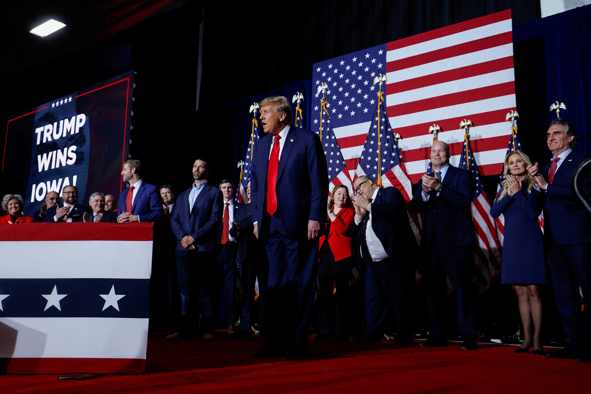 Donald Trump stands on stage surrounded by people clapping, and USA flags