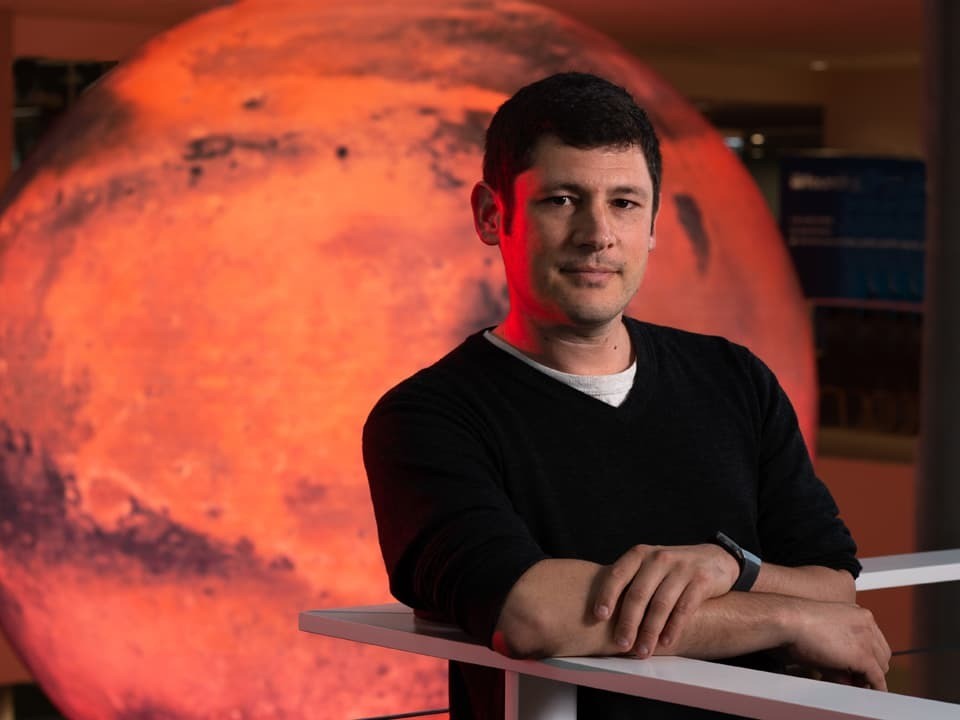 A man crosses his arms and smiles in front of a large represenation of Mars