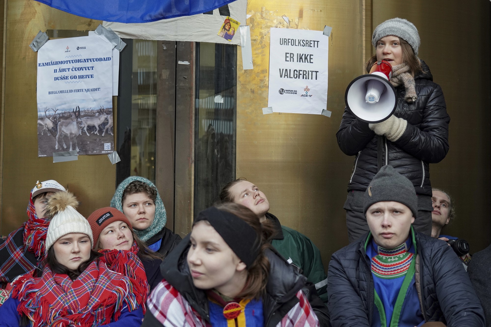 Greta Thunberg stands with a megaphone in the middle of a group of seated teenagers next to a door with taped up signs. 