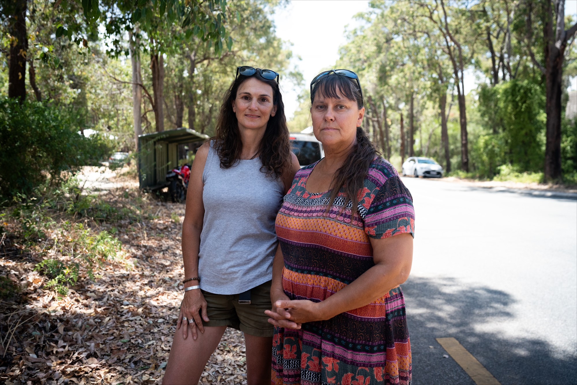 Two women stand near a road amid bushland.