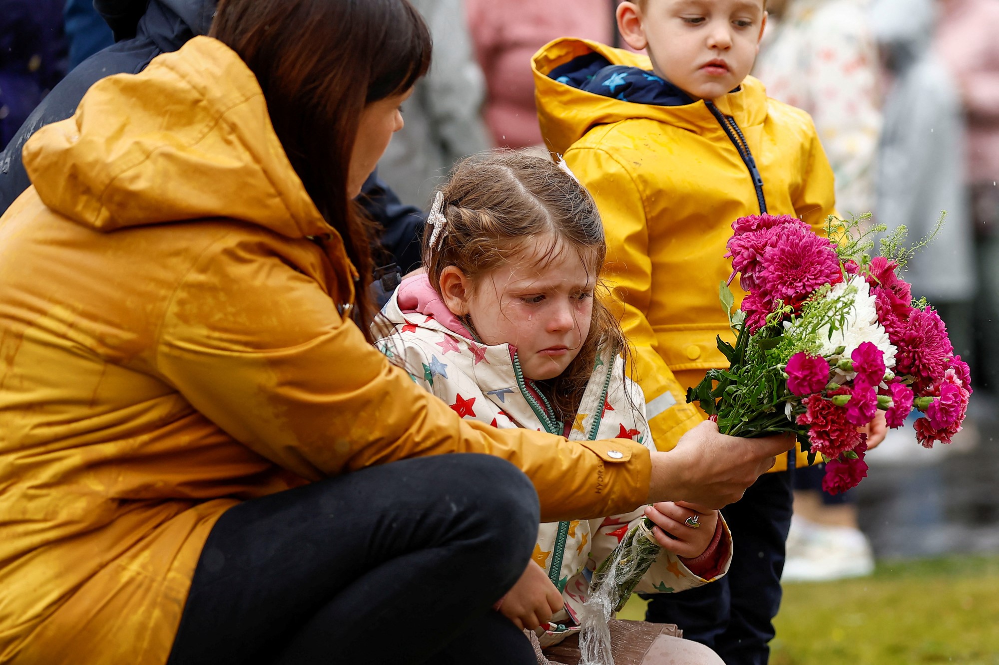A young girl is pictured with tears in her eyes, putting down flowers with a lady wearing a yellow raincoat.