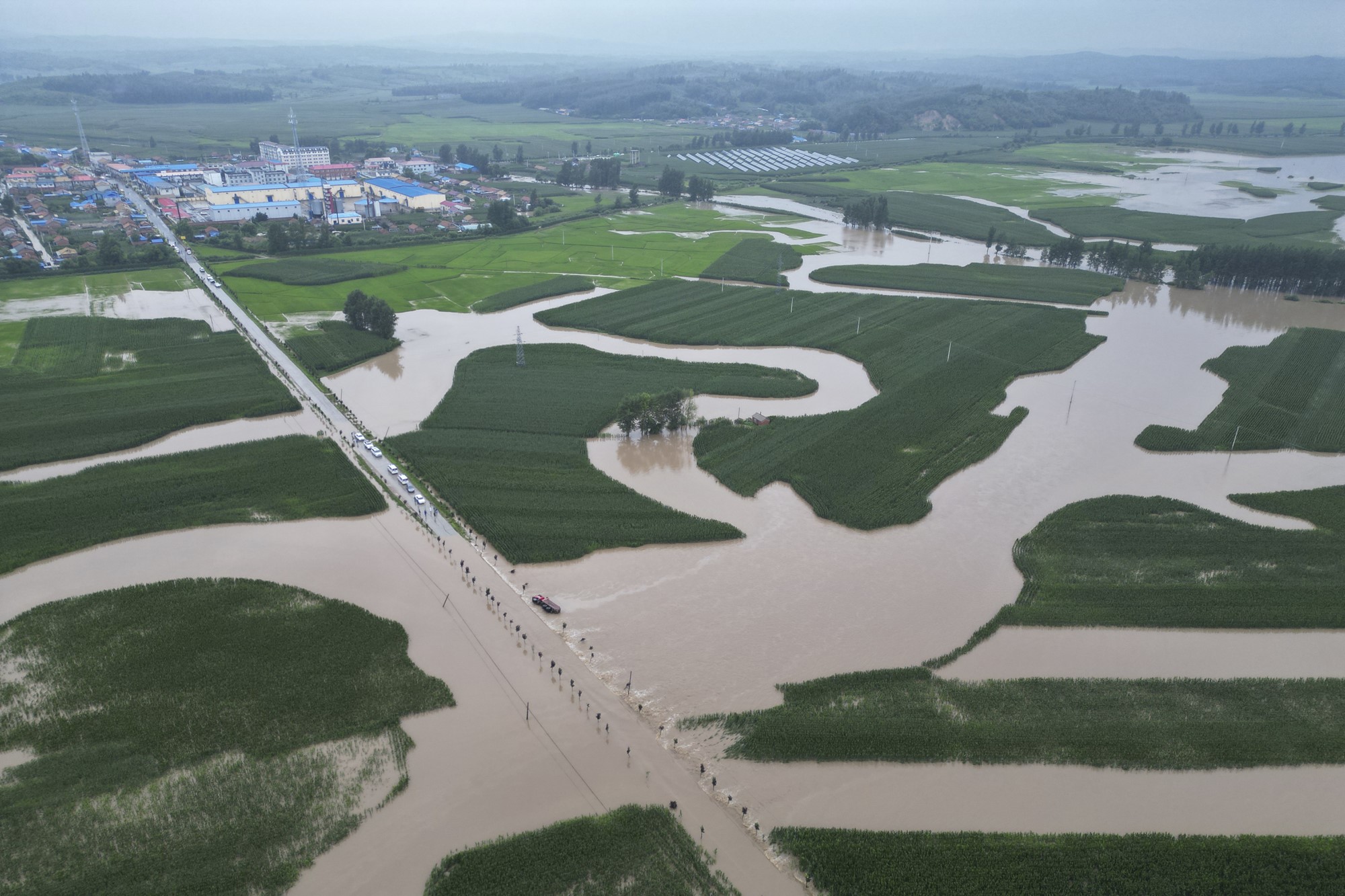 An aerial image of flooding waters over a long road and grassy plains. A town and trees in the background.