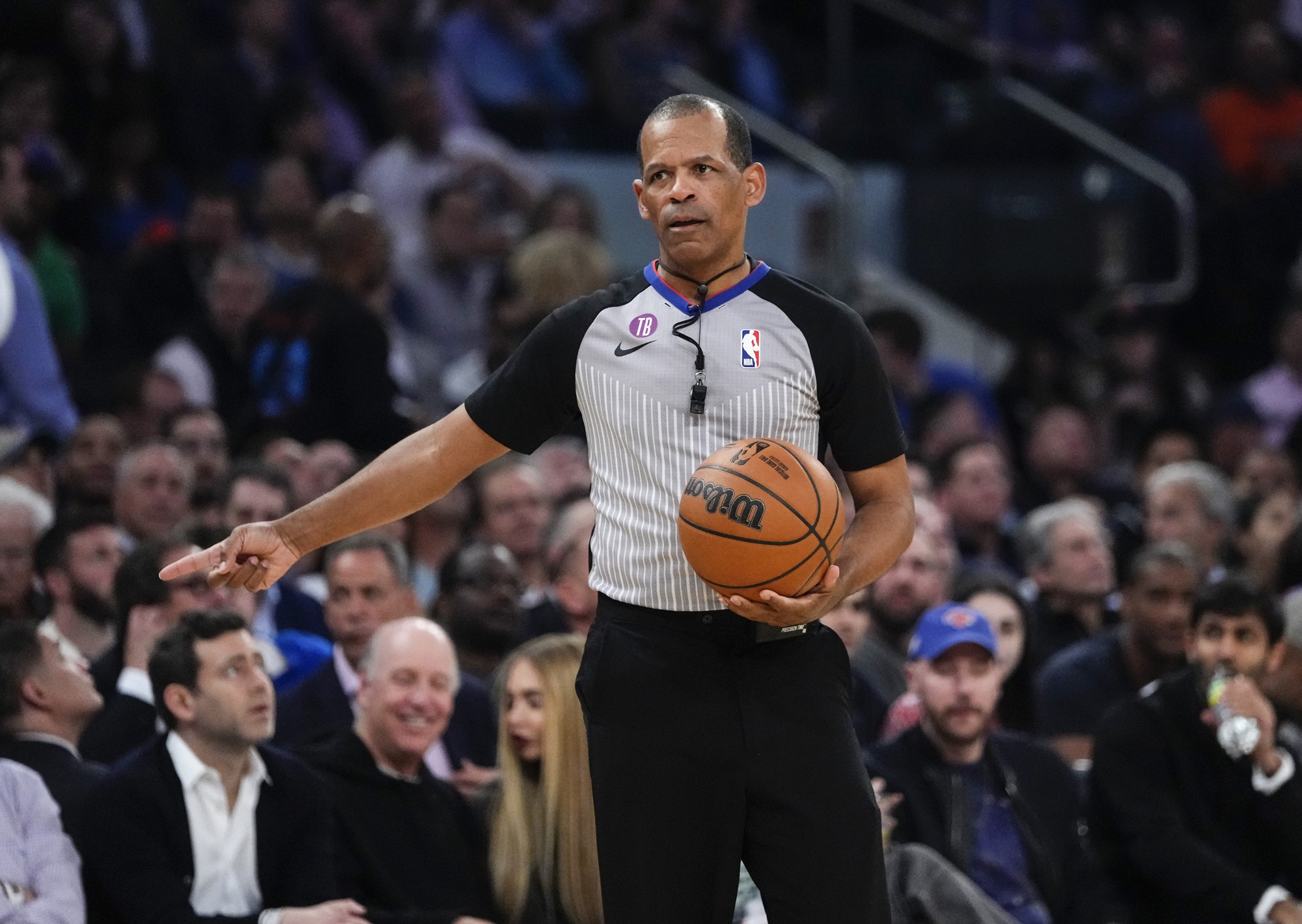 A male basketball referee holds a ball and points during a game. Crowd behind him.