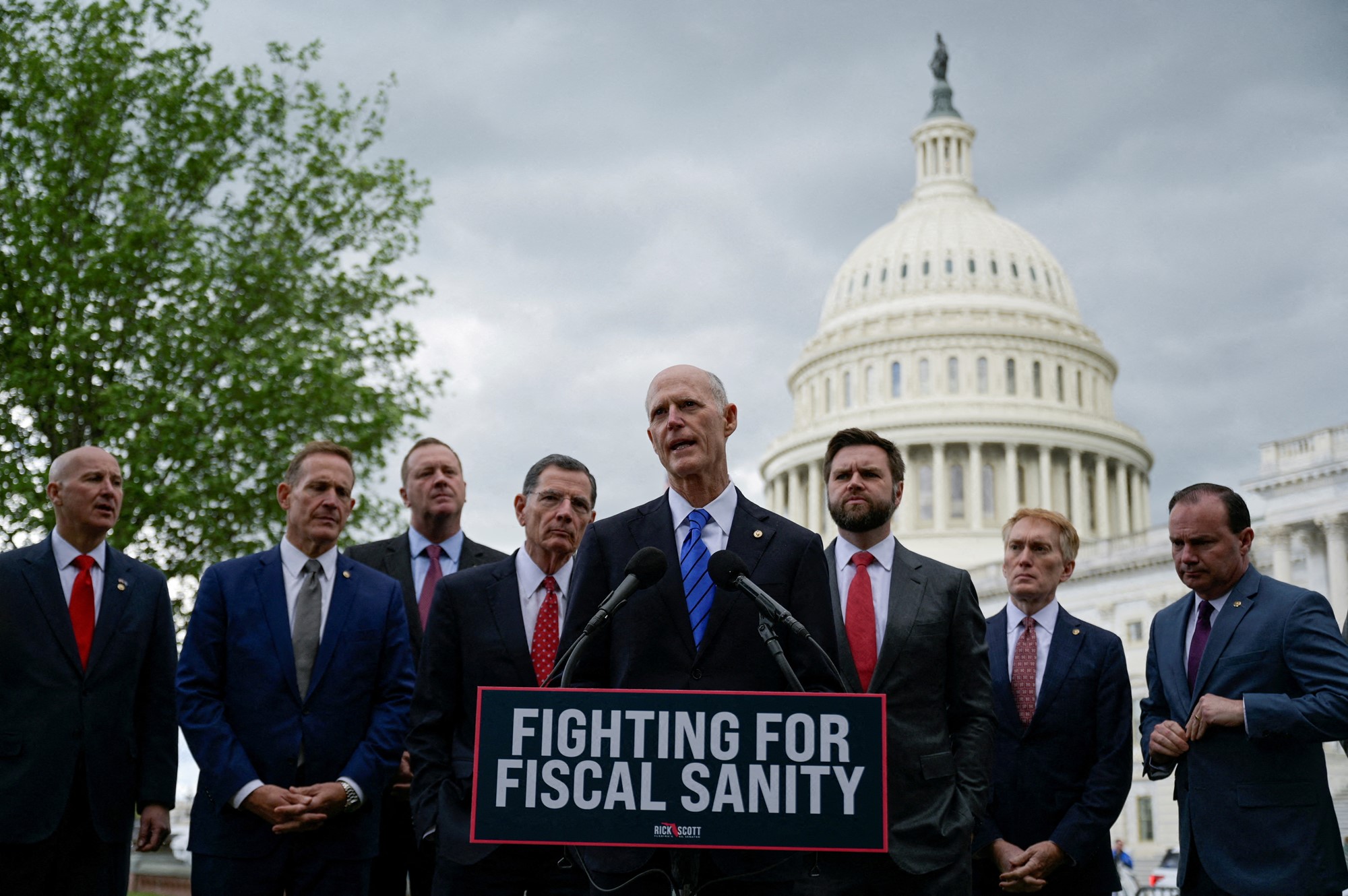 Eight white men stand behind a podium with the US Capitol building in the background.