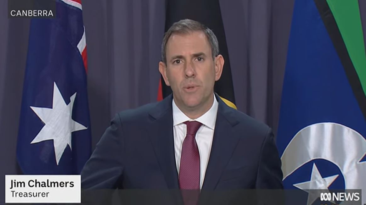 Treasurer Jim Chalmers stands in front of an Australian flag, Aboriginal flag, and Torres Strait Island flag while speaking. His name and title is superimposed on the bottom of the image, and the top left of the image has a location tag that reads "Canberra".