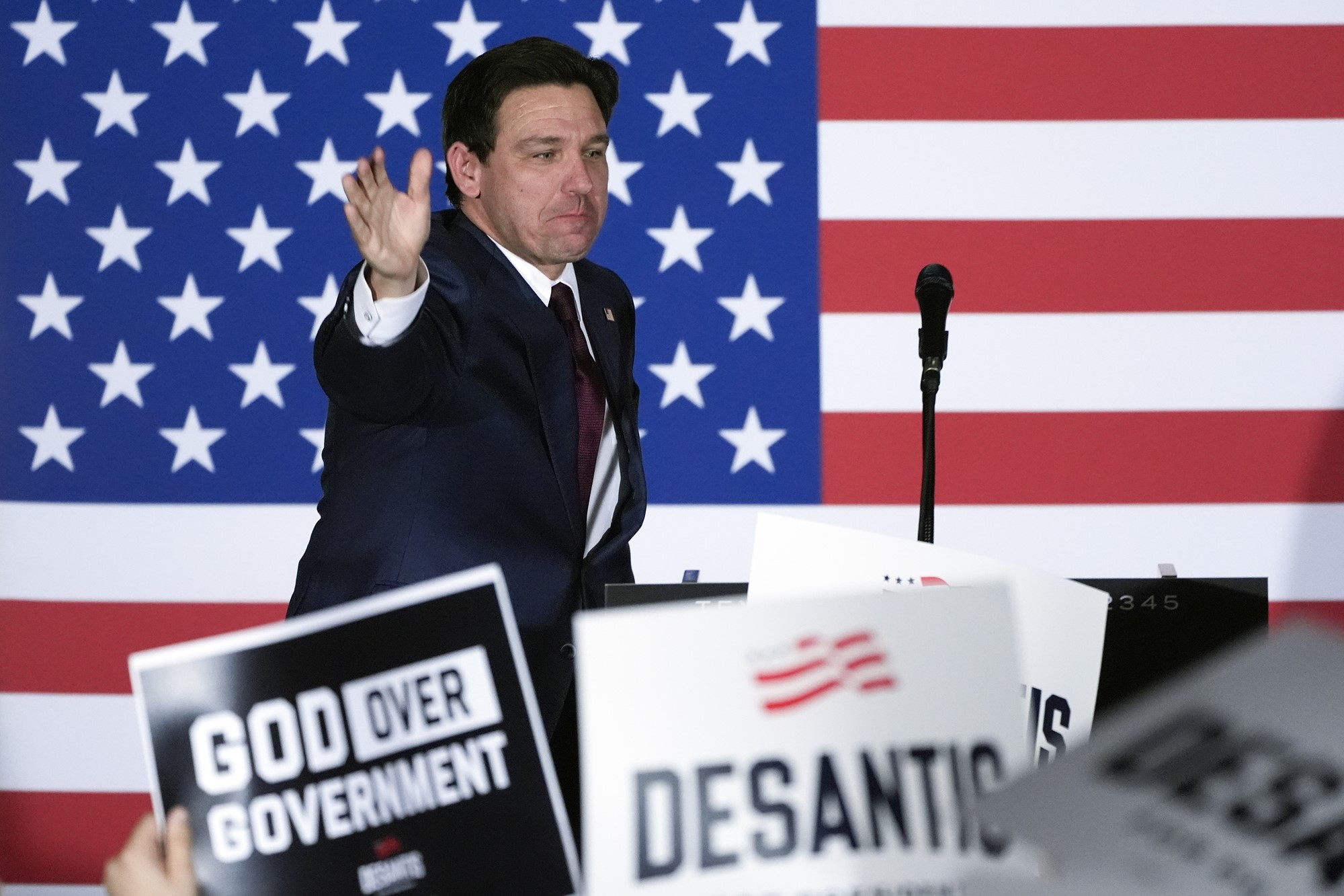 Ron Desantis on stage waving to supporters in front of a United States flag.