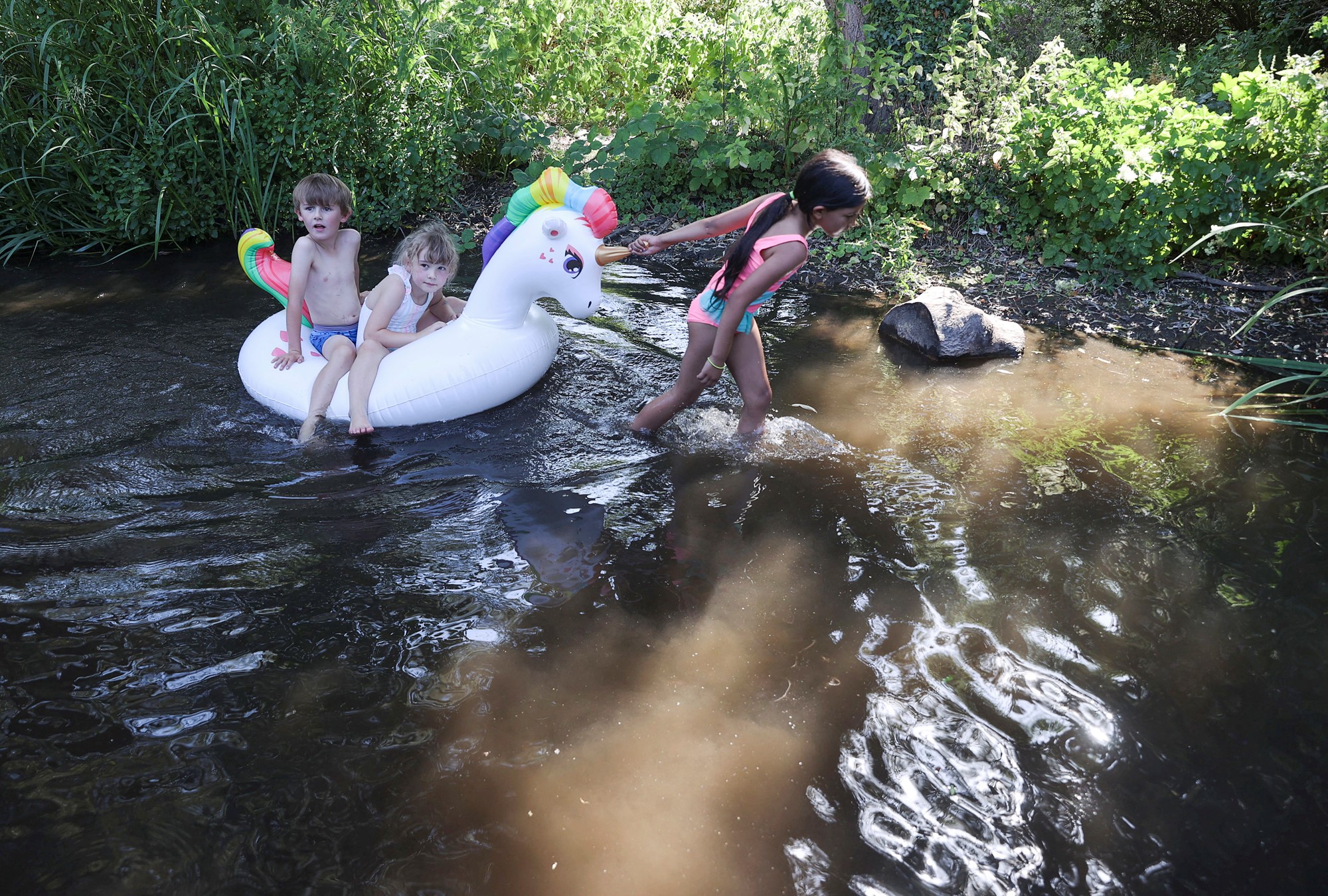 Young children play on a unicorn floating pool toy in a river.
