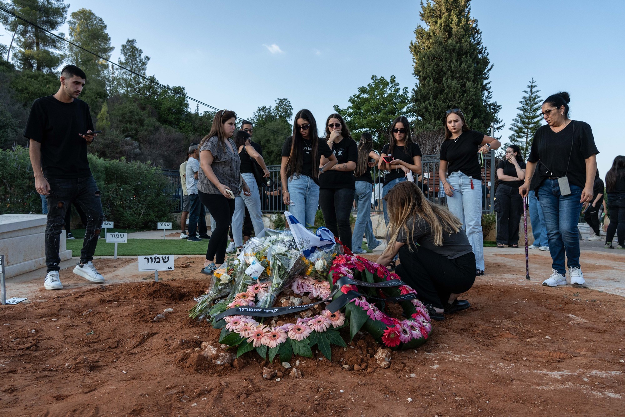 People lay wreaths at a dirt grave, as several others wearing black Tshirts and jeans watch on