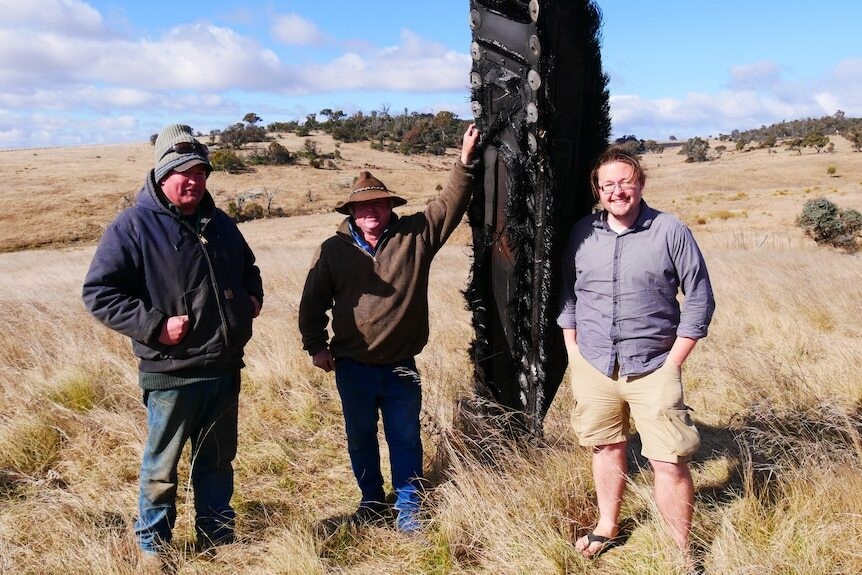 Three men stand near a large piece of space junk in a grass paddock