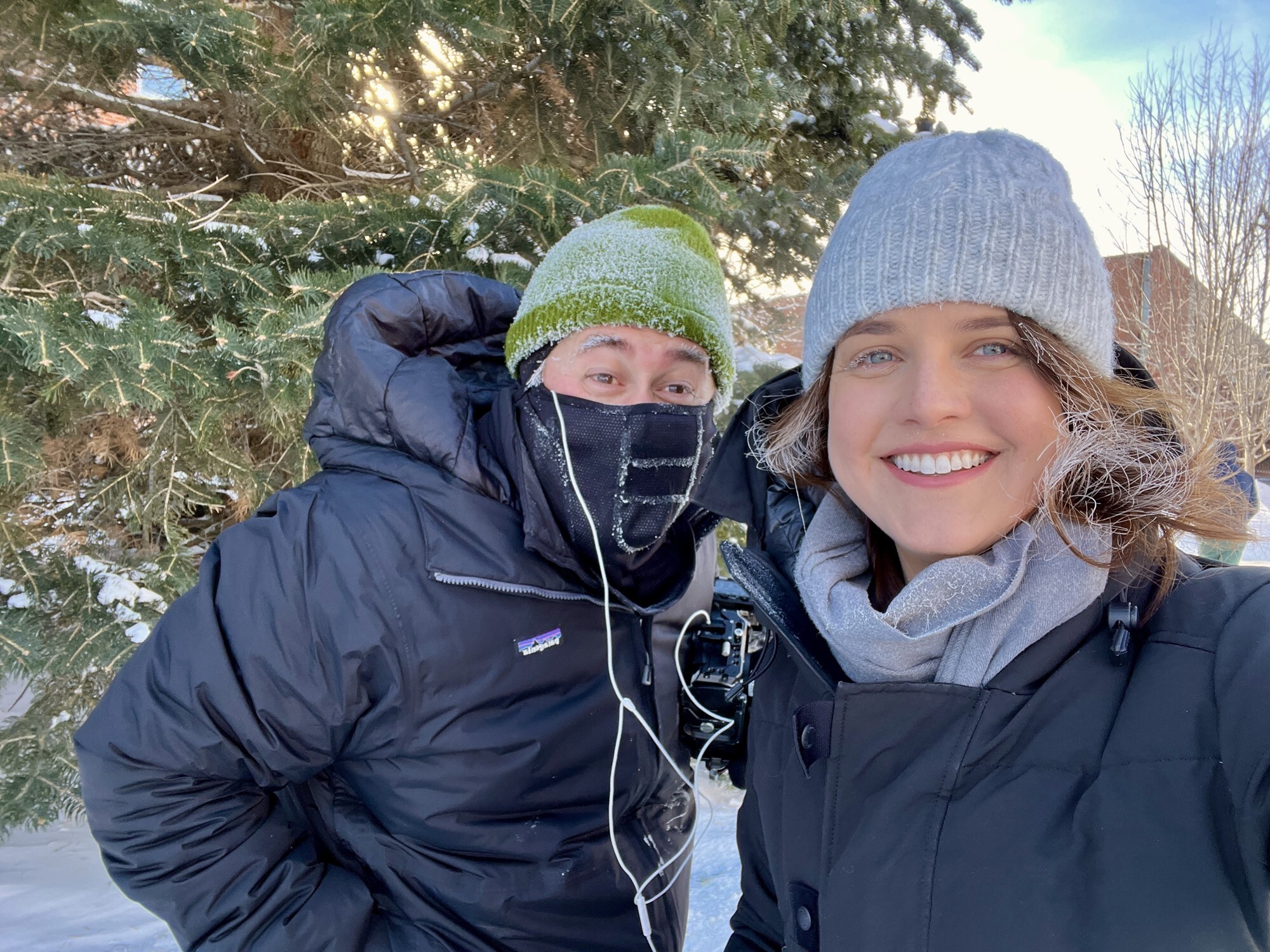 Two people in winter coats and hats, with snow in their hair and eyebrows, smile together