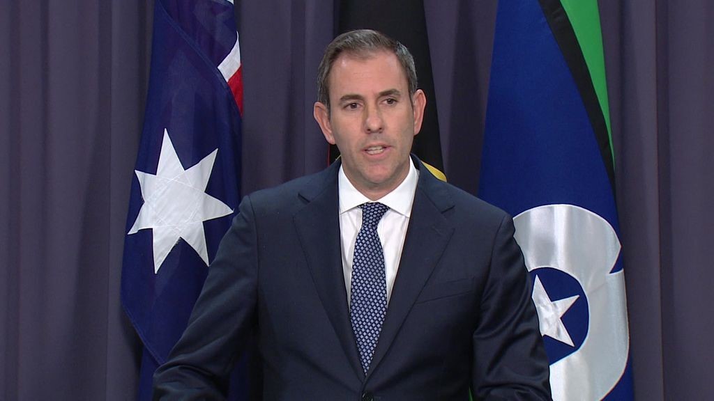 A man in a dark suit stands in front of three australian flags