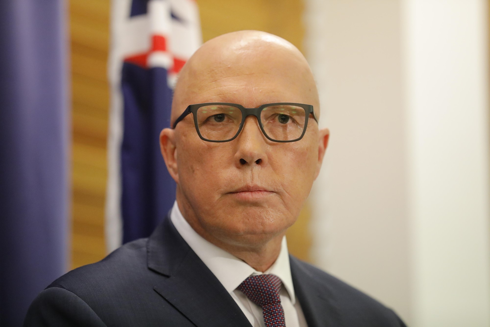 A bald man wearing glassed and a suit and tie stands in front of an Australian flag