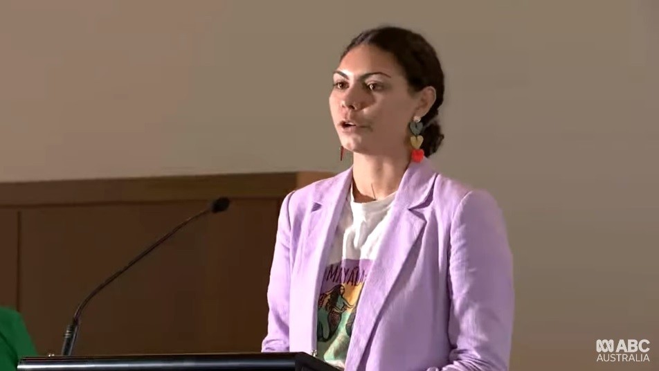 A young Indigenous woman in a pink blazer speaks at a podium.