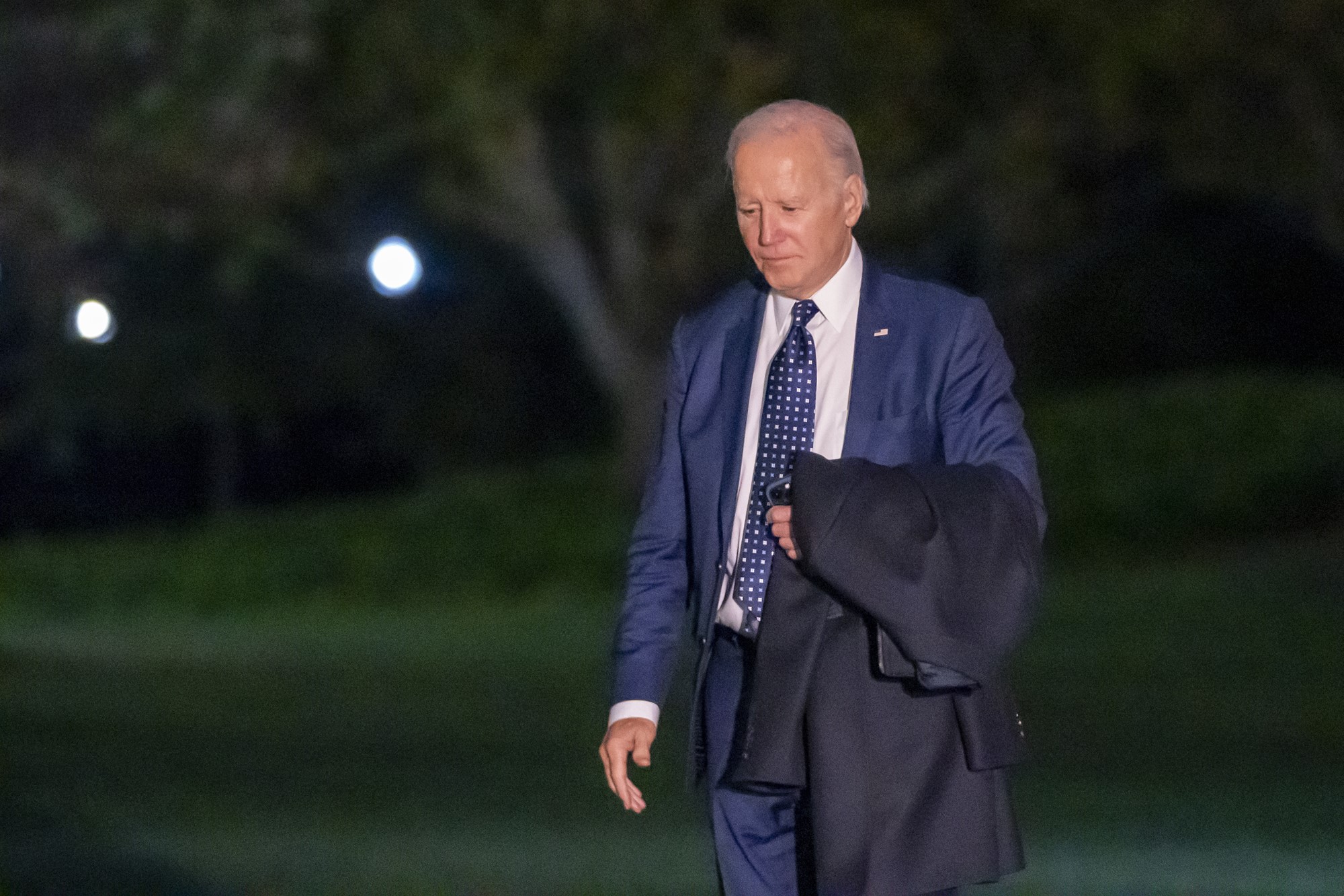 Joe Biden walks looking down, wearing a suit and carrying a jacket