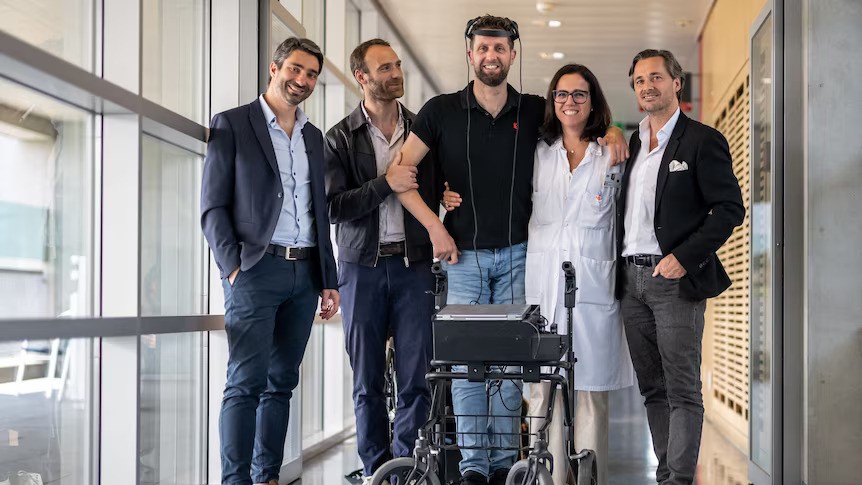 Four men and a woman embrace in the halls of a hospital, one man with a walking frame and a device with wires on his head