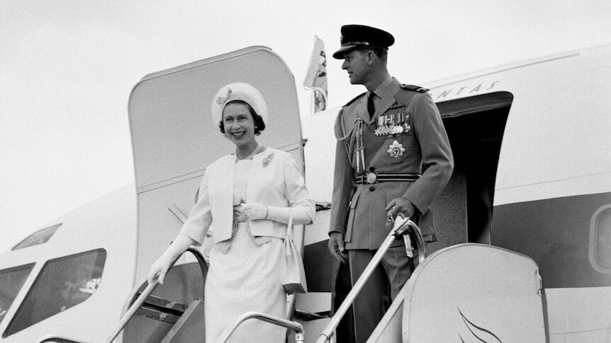 Queen Elizabeth and Prince Philip smile as they walk down the steps of a plane