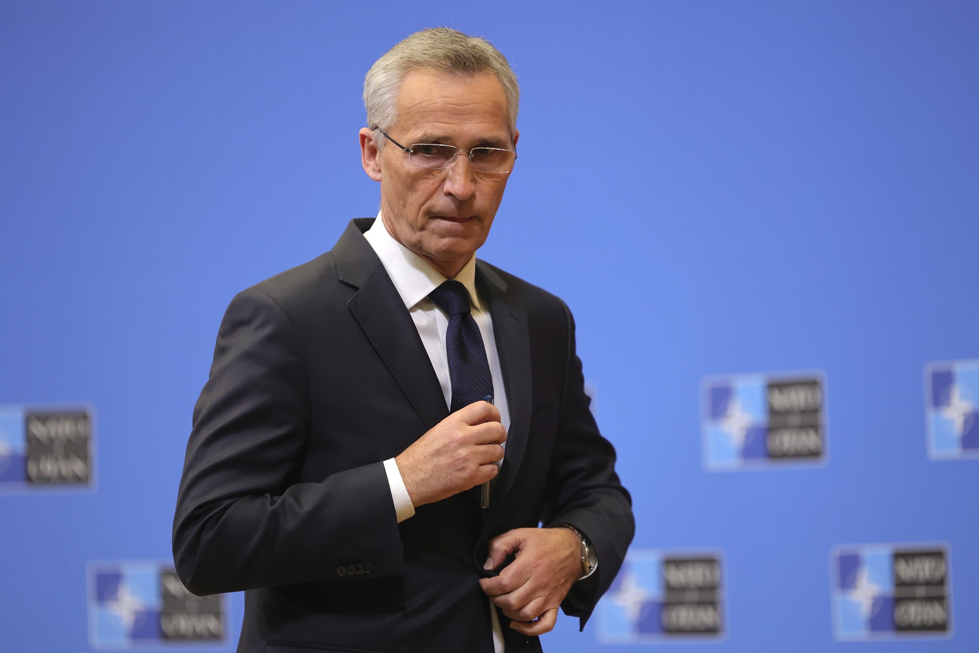 Jens Stoltenberg adjusts his suit as he looks pensively in front of a NATO backdrop