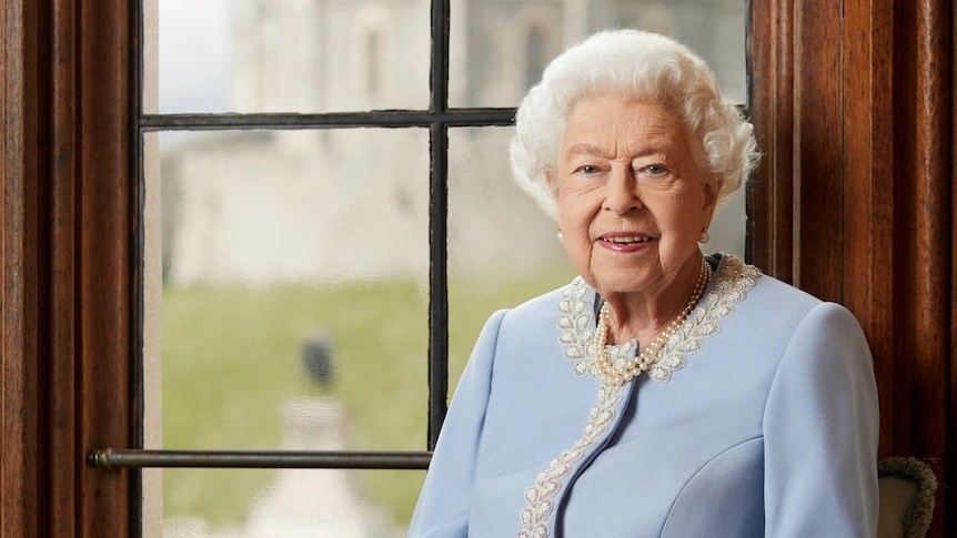 The late Queen of England dressed in a powder blue jacket with lace trim and a pearl necklace sits near a window with a castle and grass in the background.