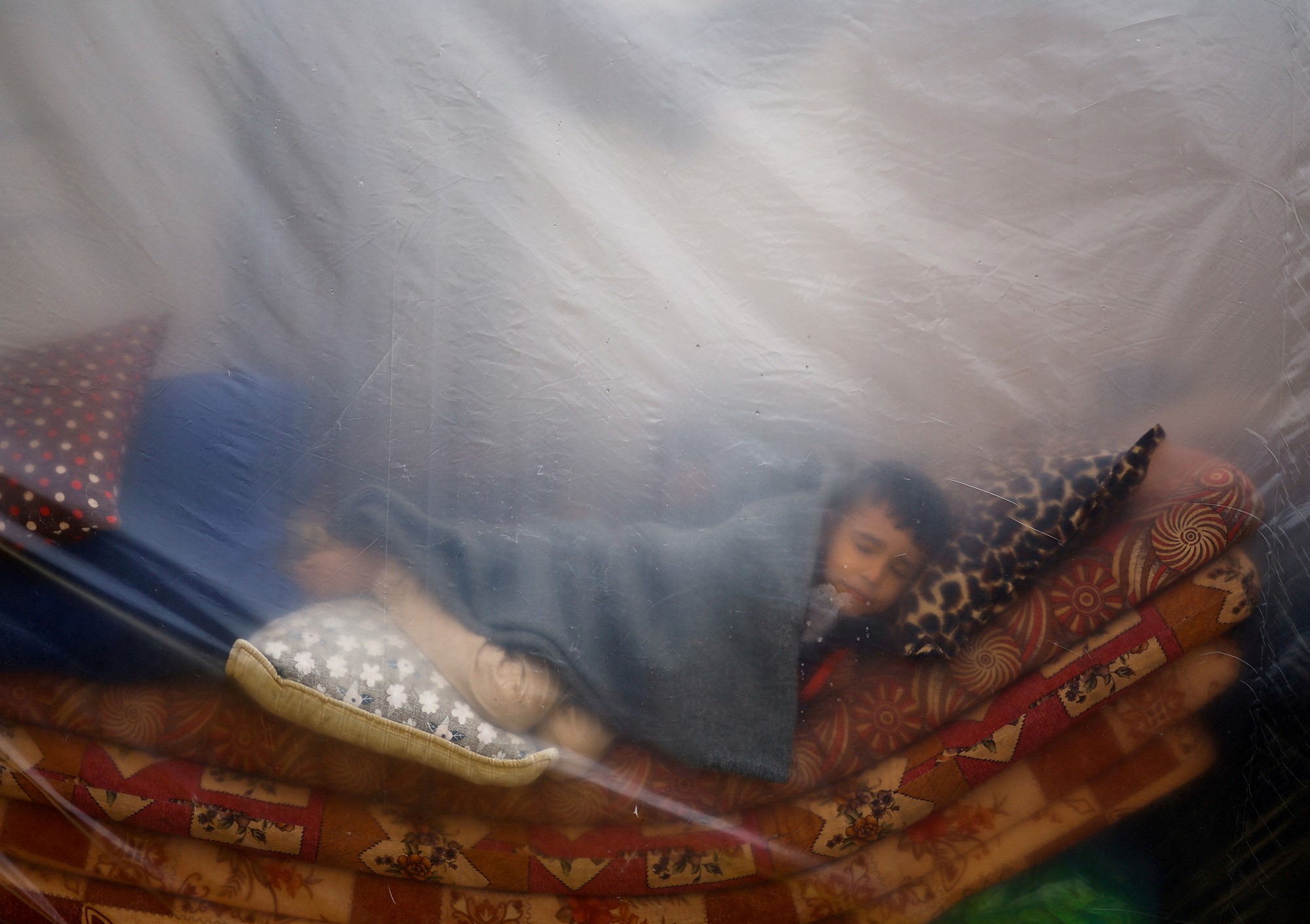 Boy sleeps on bundle of thin mattresses behind plastic sheet 