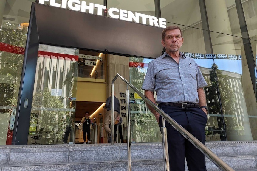 A man wearing a short collared button down shirt and black slacks stands in front of a Flight Centre office.