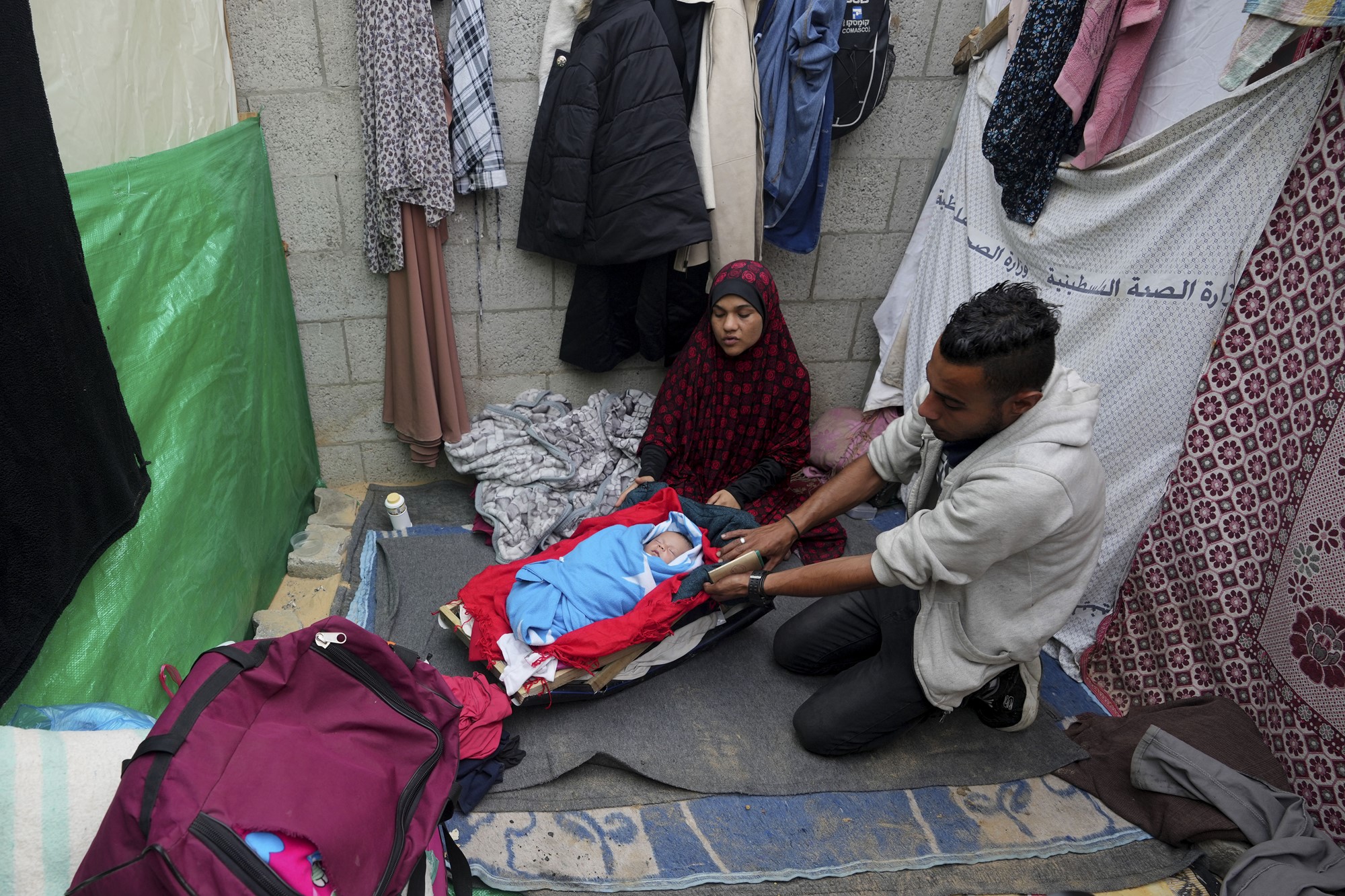 Family sits on the ground around a baby sleeping in a blue blanket. Clothes hang on the wall behind them.