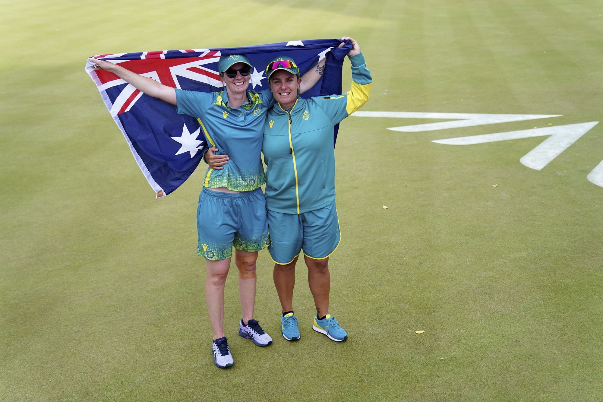 Two women in green and gold tracksuits hold an Australian flag.