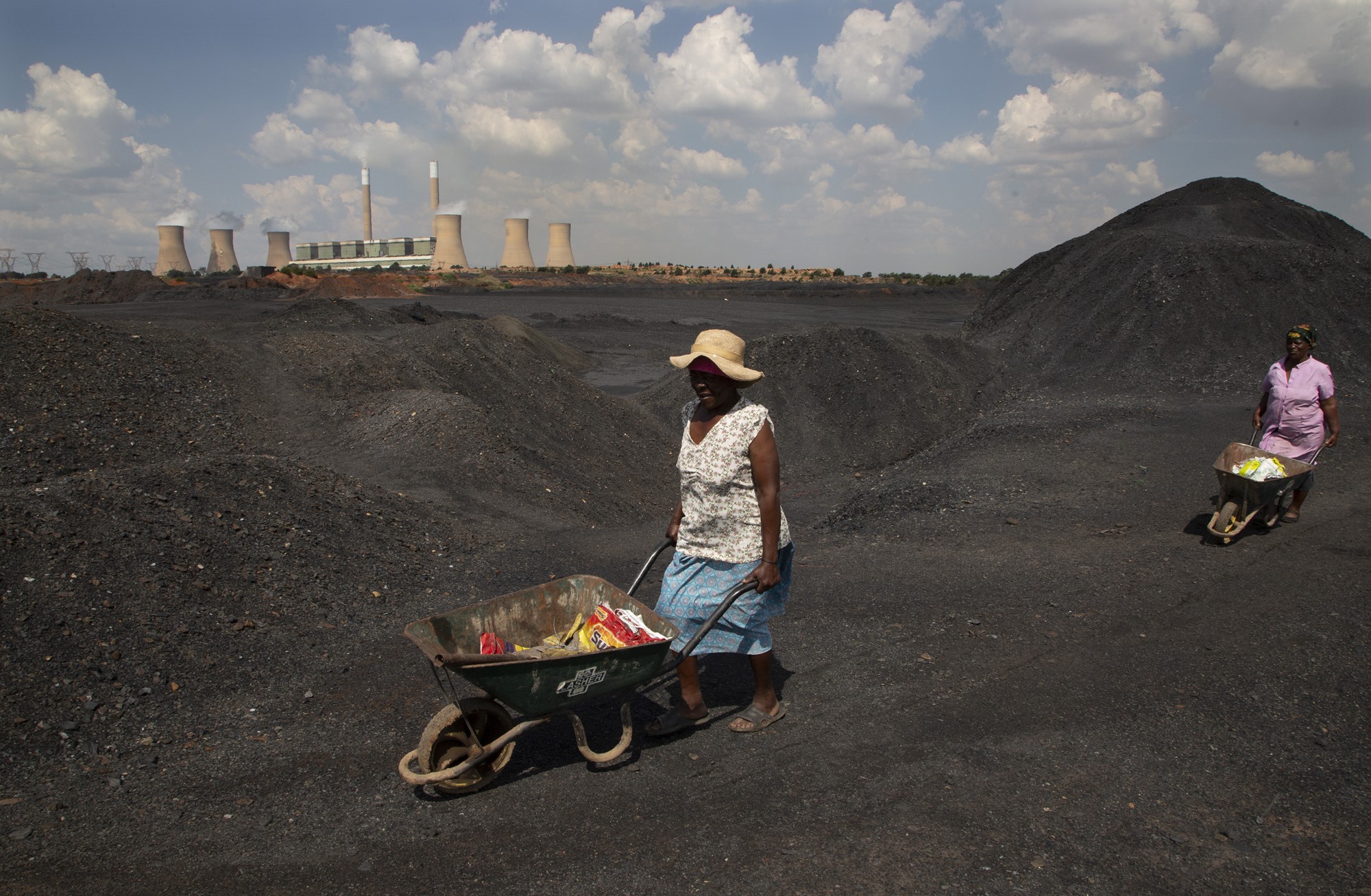 Women wearing hats push wheelbarrows over coal. 