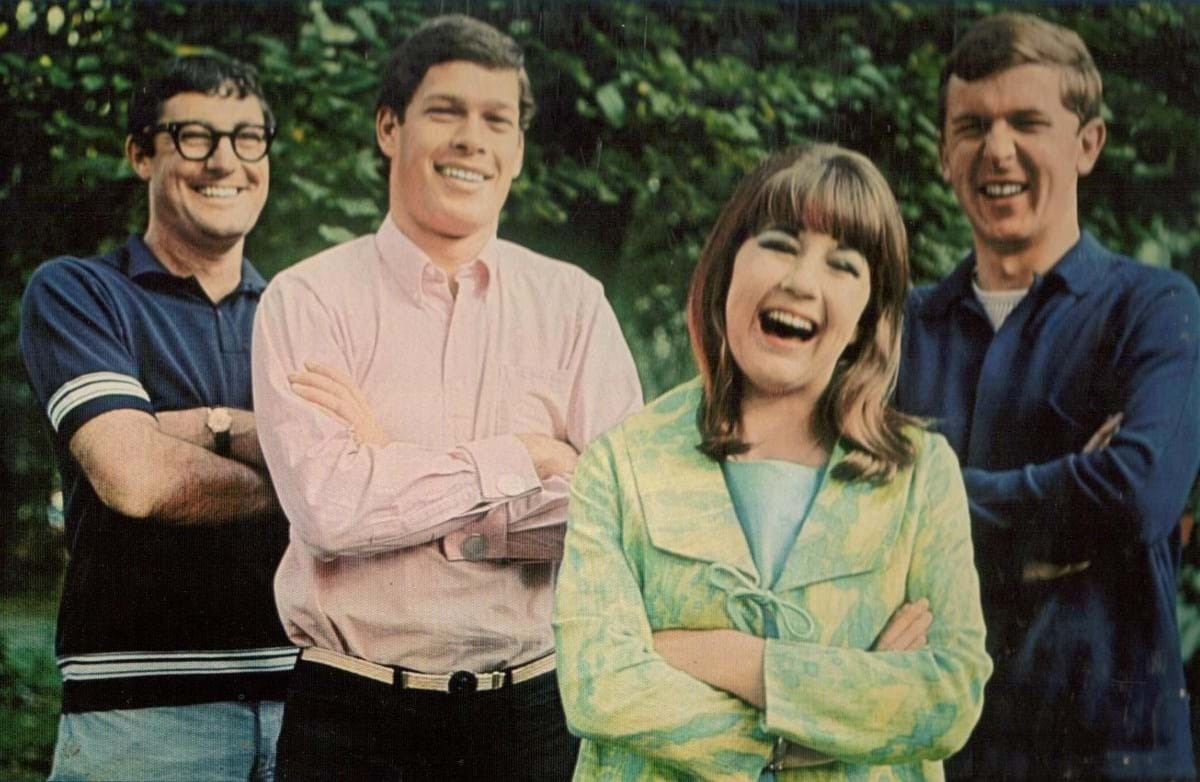 The members of The Seekers laugh, arms crossed, as they stand in a garden.