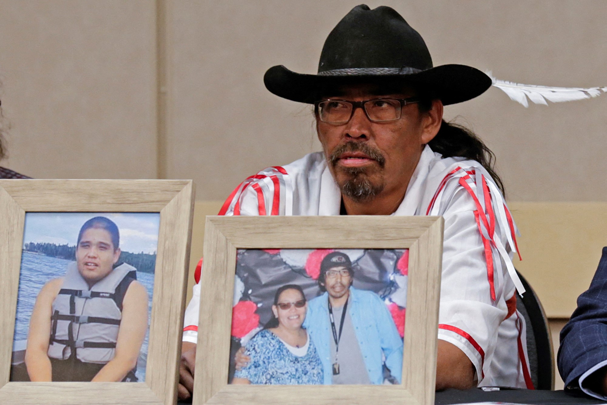 a man wearing a black hat sits at a desk behind framed photographs of stabbing victims