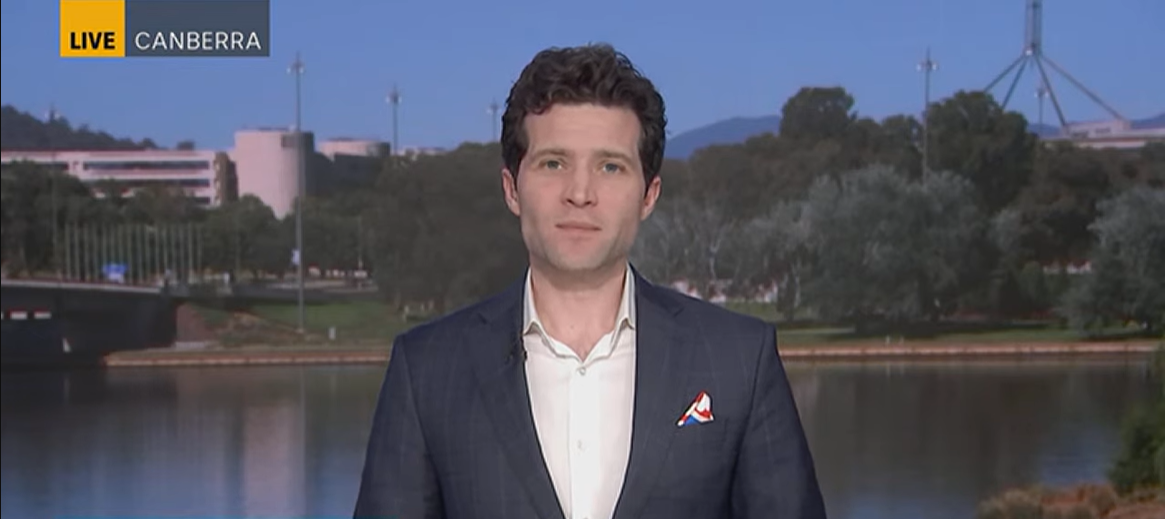 A white man in a suit gives a TV interview in front of a Canberra backdrop showing trees and a body of water near parliament house