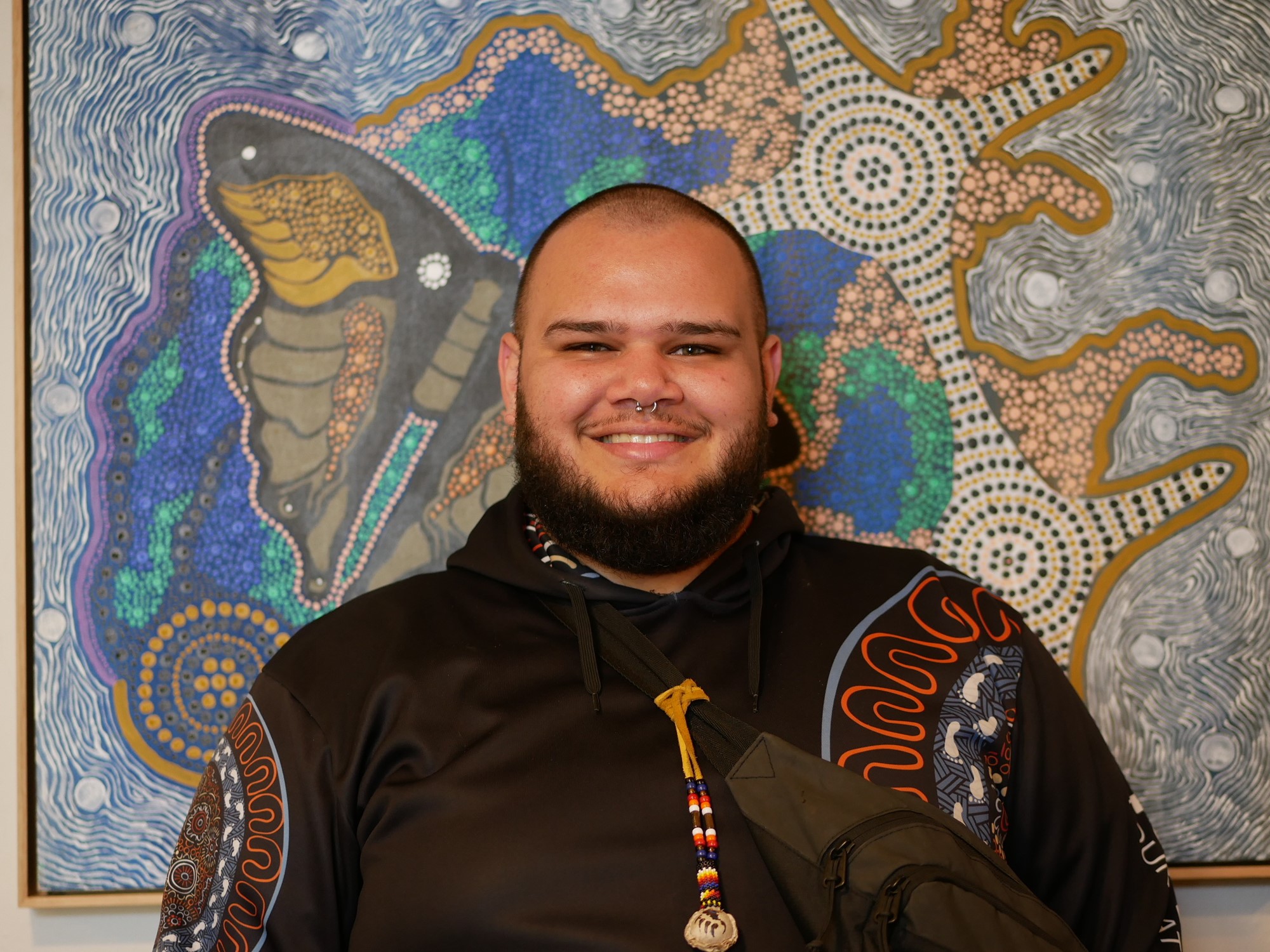 A young Aboriginal man with a nose ring stands in front of an Aboriginal painting.