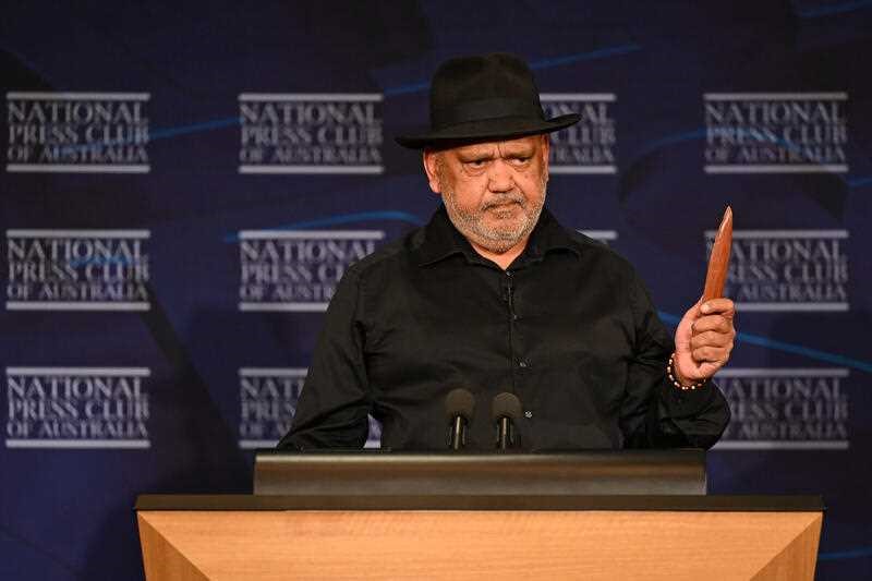 Pearson holds up an Indigenous clap stick while speaking at the podium.