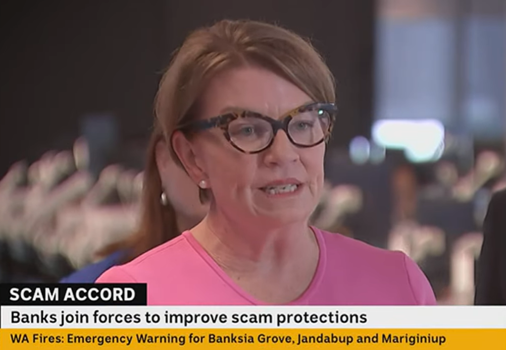 A woman with short brown hair and large cat-eye tortoiseshell glasses wearing a pink shirt is captured while talking in an unidentifiable office space. A news strap is superimposed on the bottom of the image explaining that she is speaking about banks joining forces to improve scam protections.
