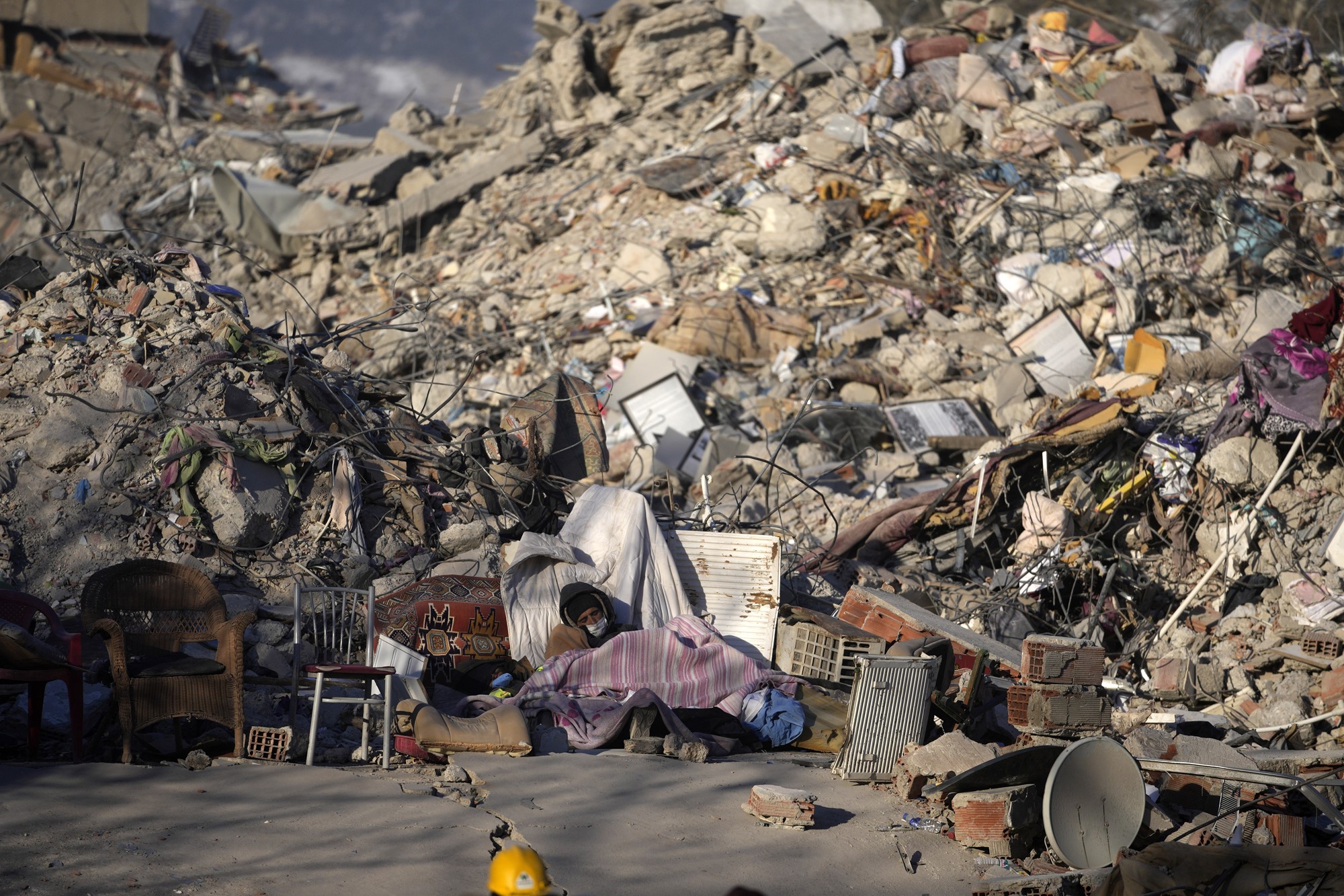 A person sleeps on the ground in front of a pile of rubble. 