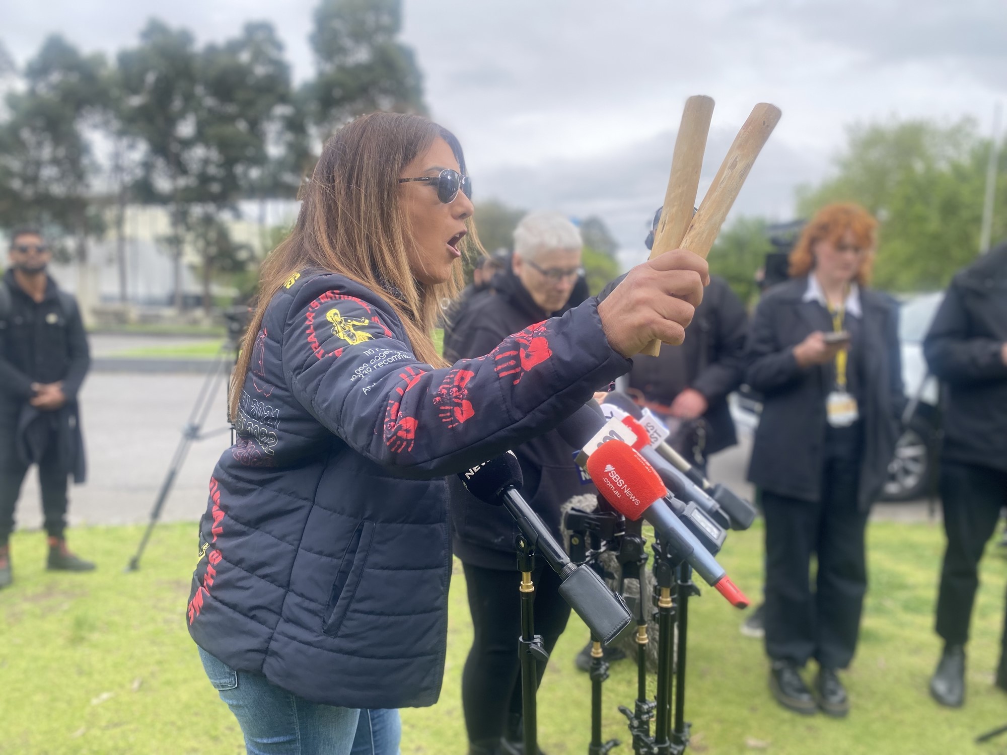 A woman holds two sticks while speaking into several microphones.