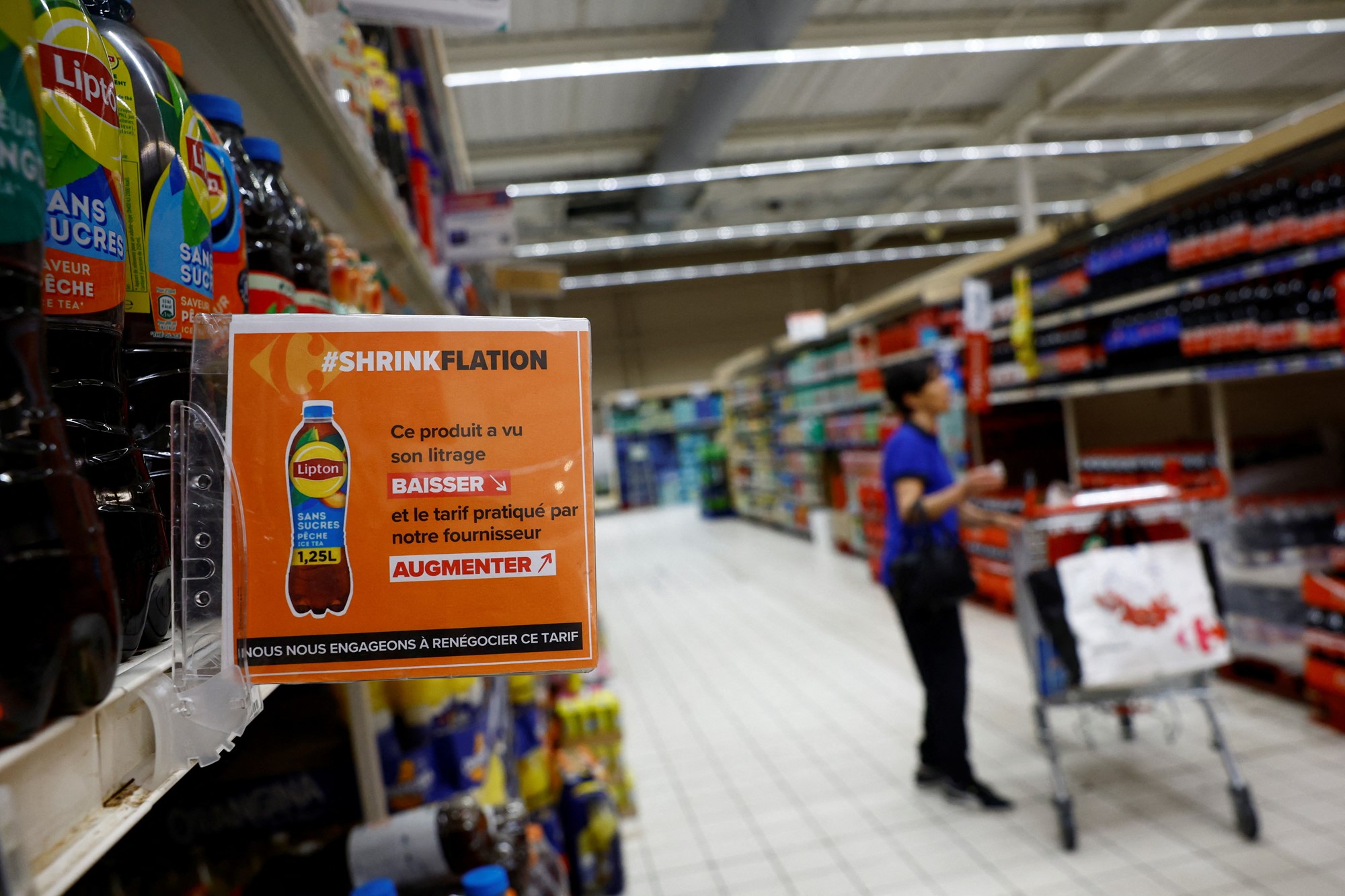 A person stands in a supermarket beverage aisle with a trolley, with a sign poking out saying #ShrinkFlation, pointing out to customers that prices have been going up while the products have shrunk in size.
