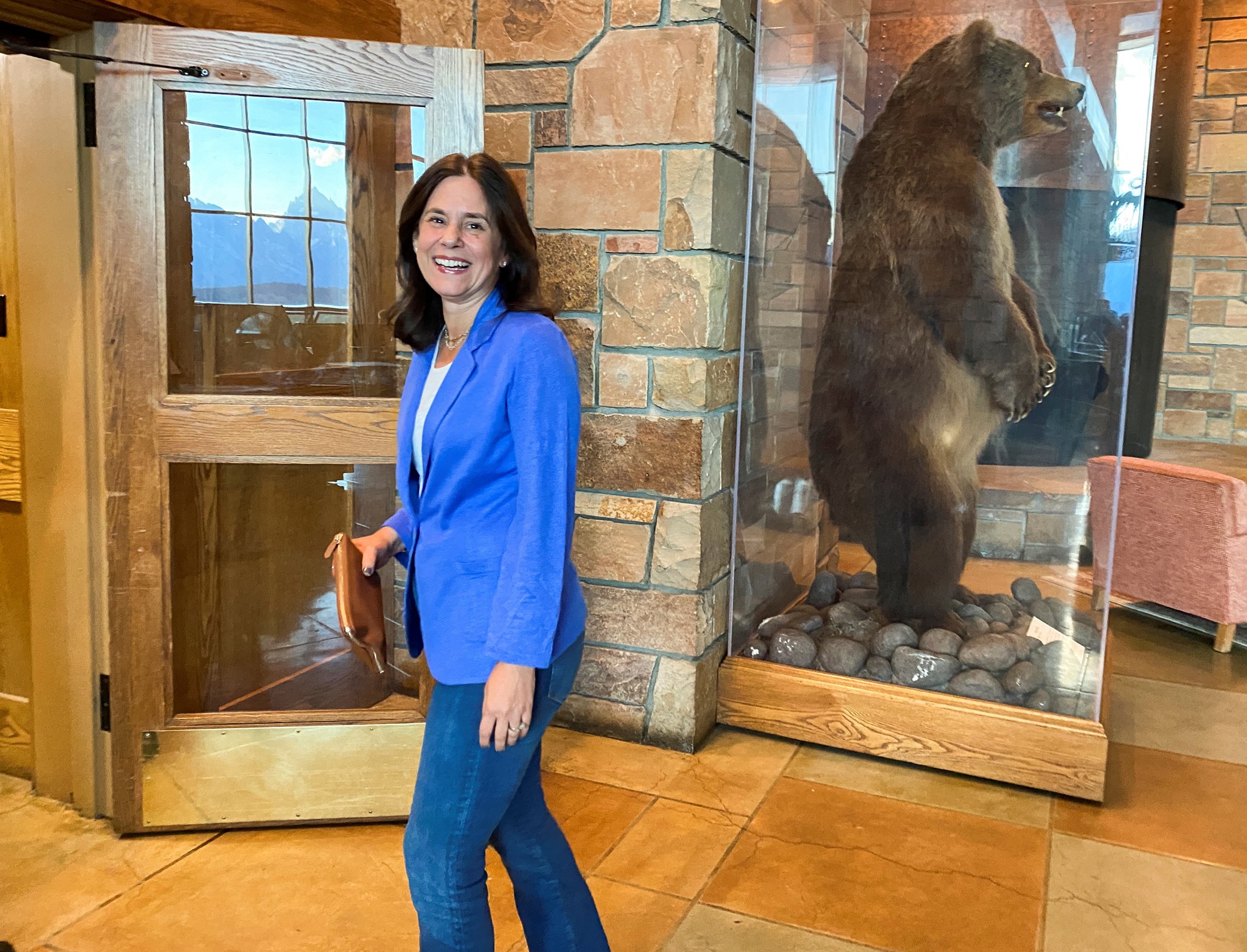 A woman wearing blue jeans and a blue blazer walks past a taxidermied bear in a lodge.