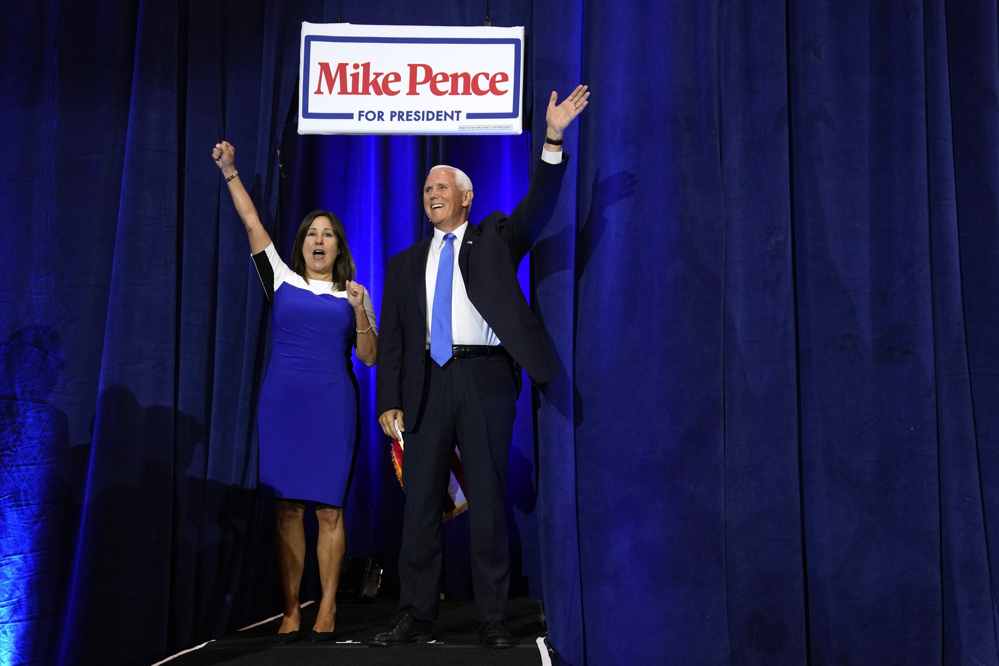 A white man stands in a suit wearing a blue tie, next to a woman in a blue dress with their hands in the air.